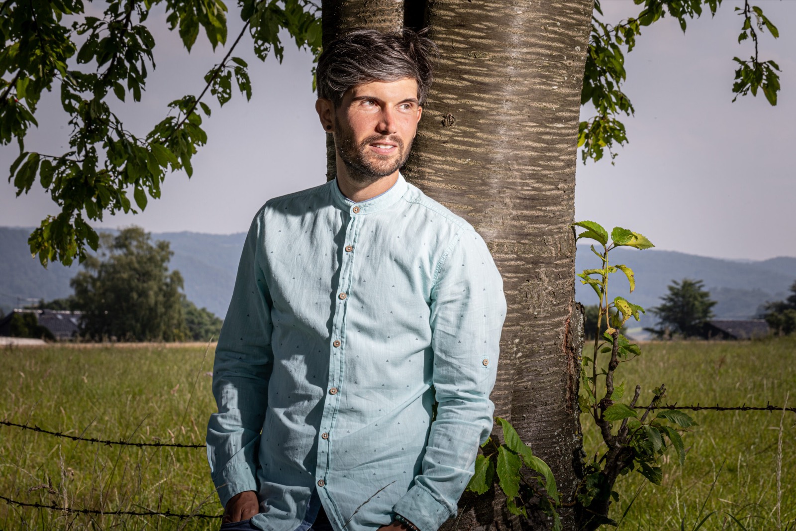 Marcus Hild lehnt an einem Baum, im Hintergrund Wiesen und Hügel des Westerwalds.