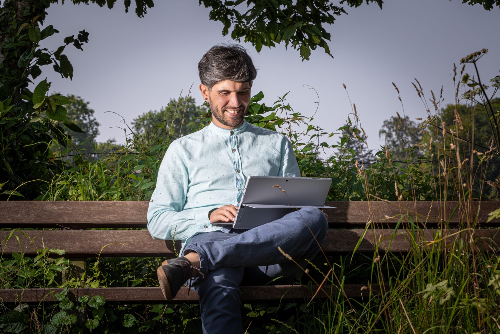 Marcus Hild sitzt lachend mit einem Laptop auf einer Holzbank im Grünen.
